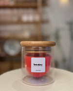 A red candle inside a double glass jar with a wooden lid, labeled 'love story' on a white surface with blurred shelves in the background.