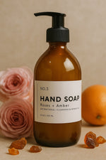 Hand soap bottle labeled 'Roses + Amber' with flowers and fruits on a beige background