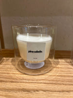 A candle with a label that reads 'Pina Colada' in a transparent double-walled glass holder, with the candle wax visible and a wooden surface in the background.