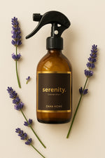 A brown glass bottle with a black spray nozzle labeled 'Serenity' by Zaha Home, surrounded by lavender flowers.
