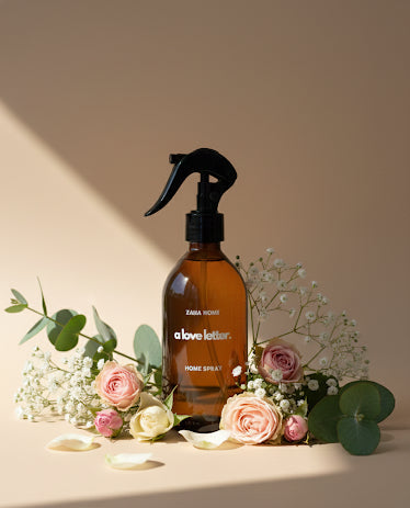 Brown spray bottle with black nozzle on a light surface surrounded by flowers and greenery, with 'zahahome.com' branding.