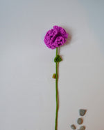A handmade crochet purple flower with a green stem and leaves, displayed against a white background with a few stones to the side.