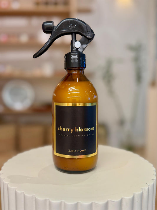 A room and fabric spray bottle with a cherry blossom label and a black spray nozzle, placed on a white surface.