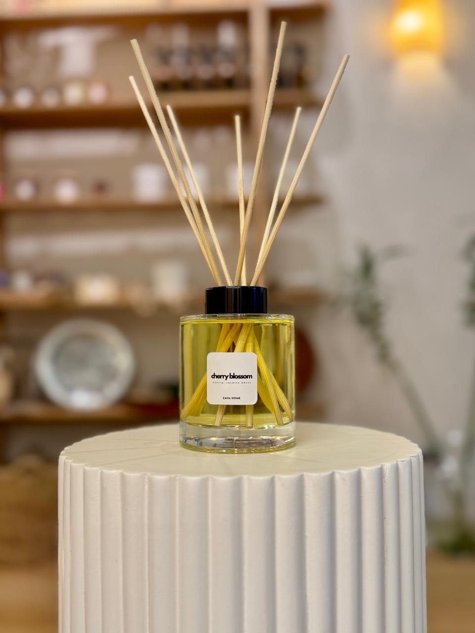 A reed diffuser with Royal Oud fragrance, featuring clear glass bottle and multiple wooden reeds, placed on a white column with a blurred background.