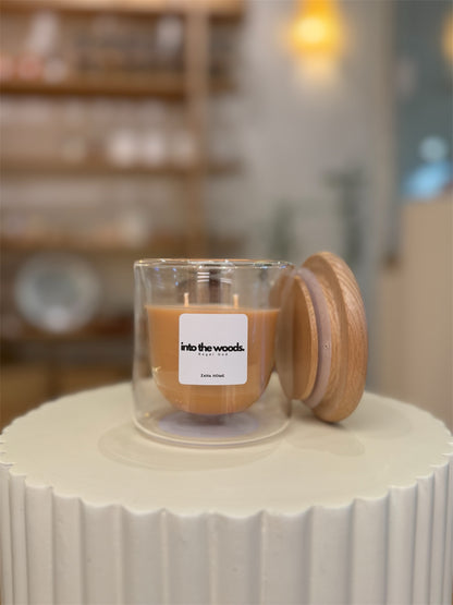 A candle with a wooden lid, glass body, and a label that reads 'Into the Woods'. The candle is placed on a white surface with blurred objects in the background.