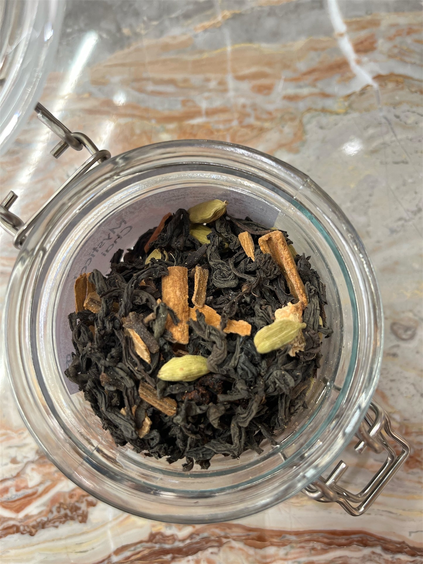 Clear glass jar with airtight lid containing black tea leaves and cardamom pods, cinnamon rolls & cloves on a marble surface.