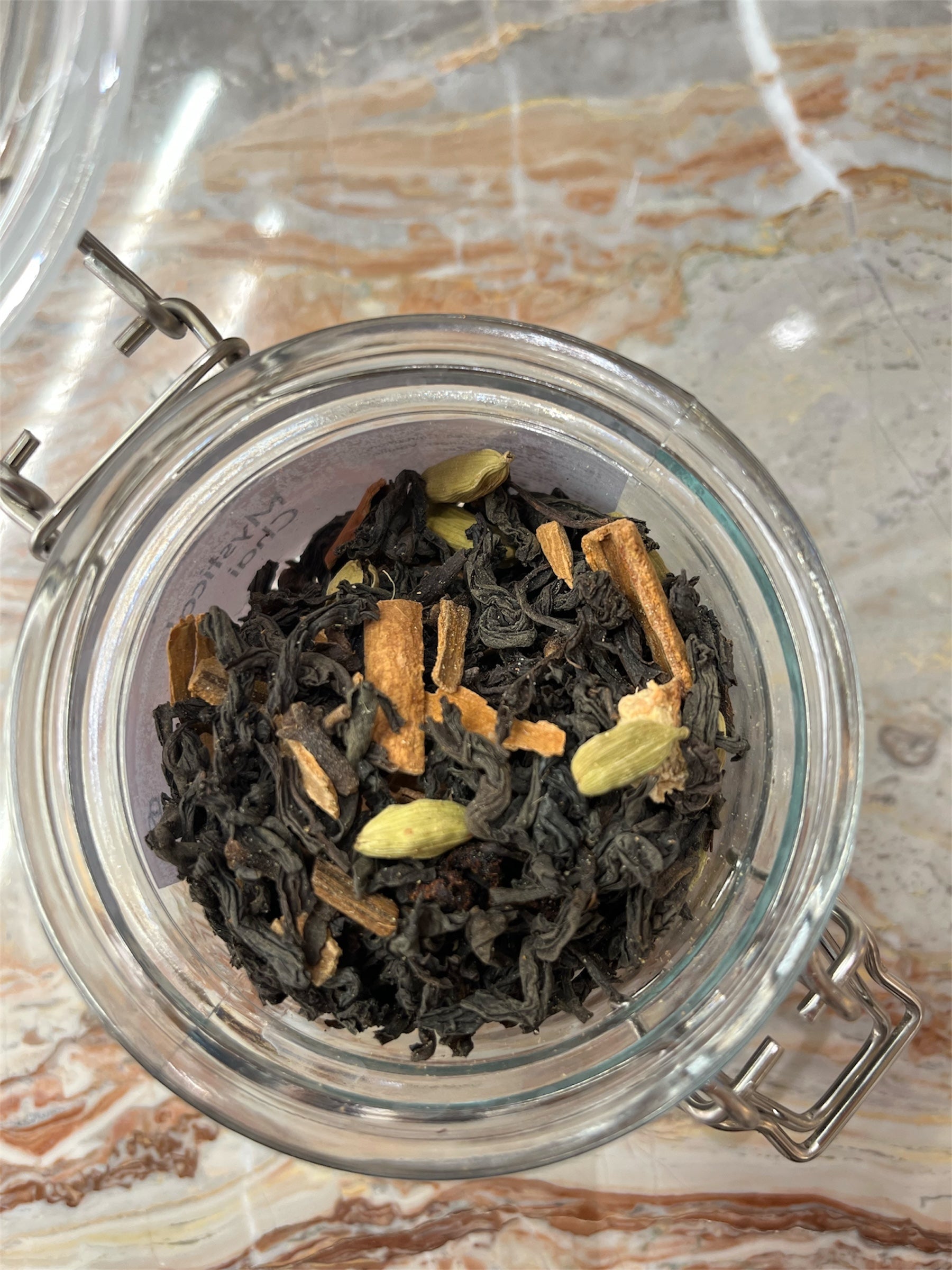 Clear glass jar with airtight lid containing black tea leaves and cardamom pods, cinnamon rolls & cloves on a marble surface.