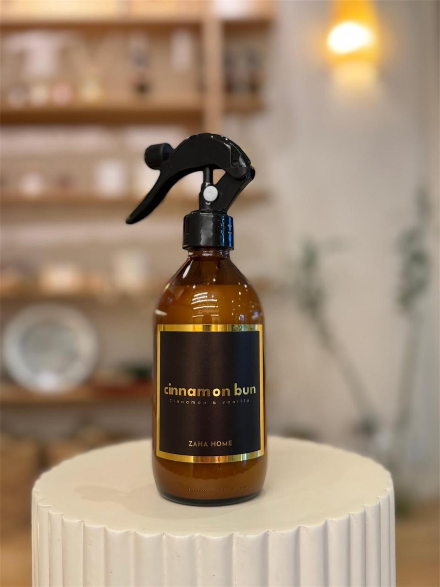 Brown spray bottle labeled 'cinnamon bun' by Zara Home on a white pedestal with a blurred indoor background.