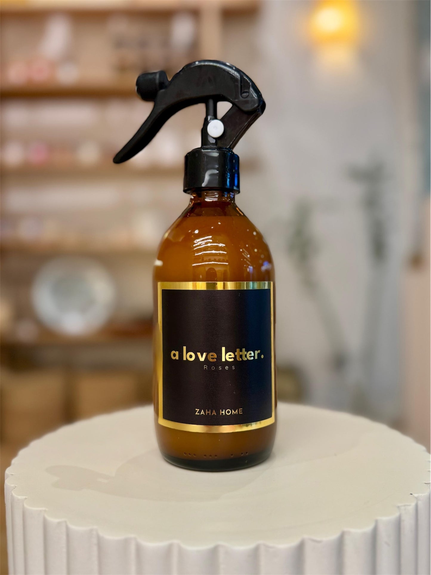A brown glass bottle with a black spray nozzle, labeled 'A Love Letter' with white and gold text, on a white surface with blurred lights and shelves in the background.
