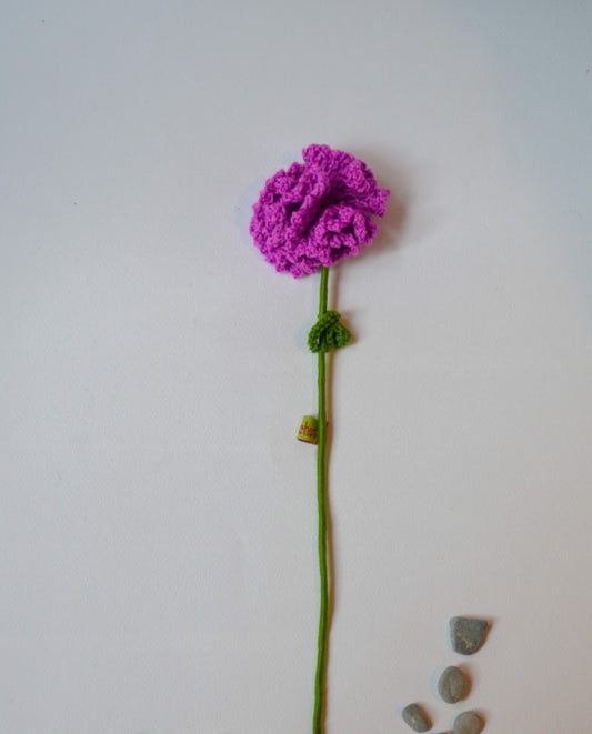 A handmade crochet purple flower with a green stem and leaves, displayed against a white background with a few stones to the side.
